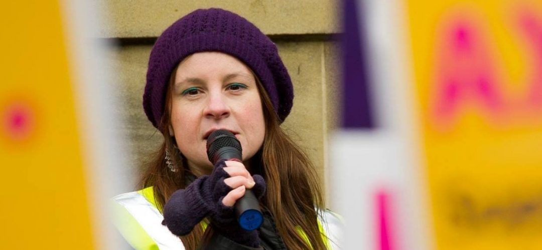 Photo of me in strike tabard (hi vis) and purple beret speaking into microphone, visible between out of focus strike placards