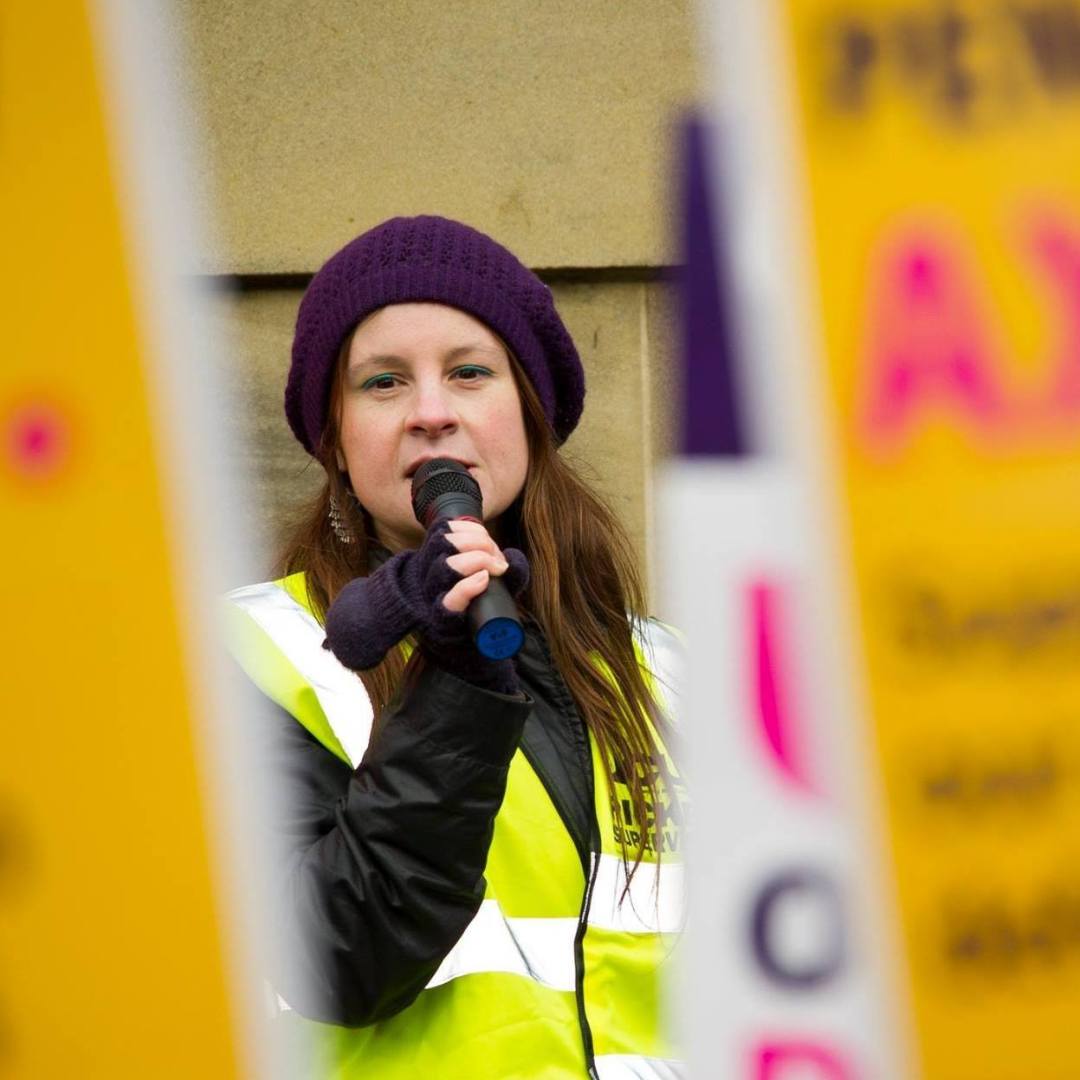 Photo of me in strike tabard (hi vis) and purple beret speaking into microphone, visible between out of focus strike placards