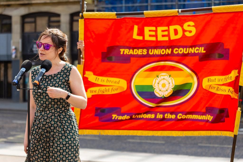 Photo is of Vicky speaking at a microphone in front of the Leeds Trades Union Council banner, which is red with yellow writing and a white Yorkshire rose against a rainbow flag.