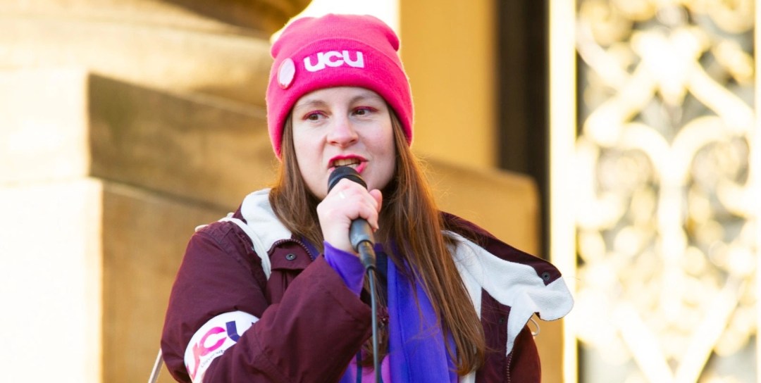 Photo of Vicky Blake wearing a plum coloured winter coat, with a UCU picket armband on, and a purple scarf with pink UCU beanie. Vicky has long brown hair and is holding a microphone speaking out to a crowd not in shot