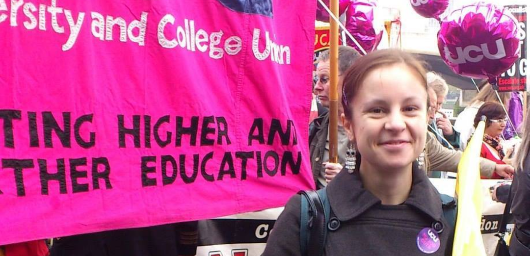 Vicky Blake smiling on the right hand side of a picture, with pink balloons that say "UCU" in white in the background where others are visible on a march. To the left is the lower right habd quarter of the UCU banner which is pink with lettering stitched on in purple and white - enbough is visible to deduce the banner says University and College Union and "--TING HIGHER AND (--THER EDUCATION" (Probably Protecting Higher and Further Education)