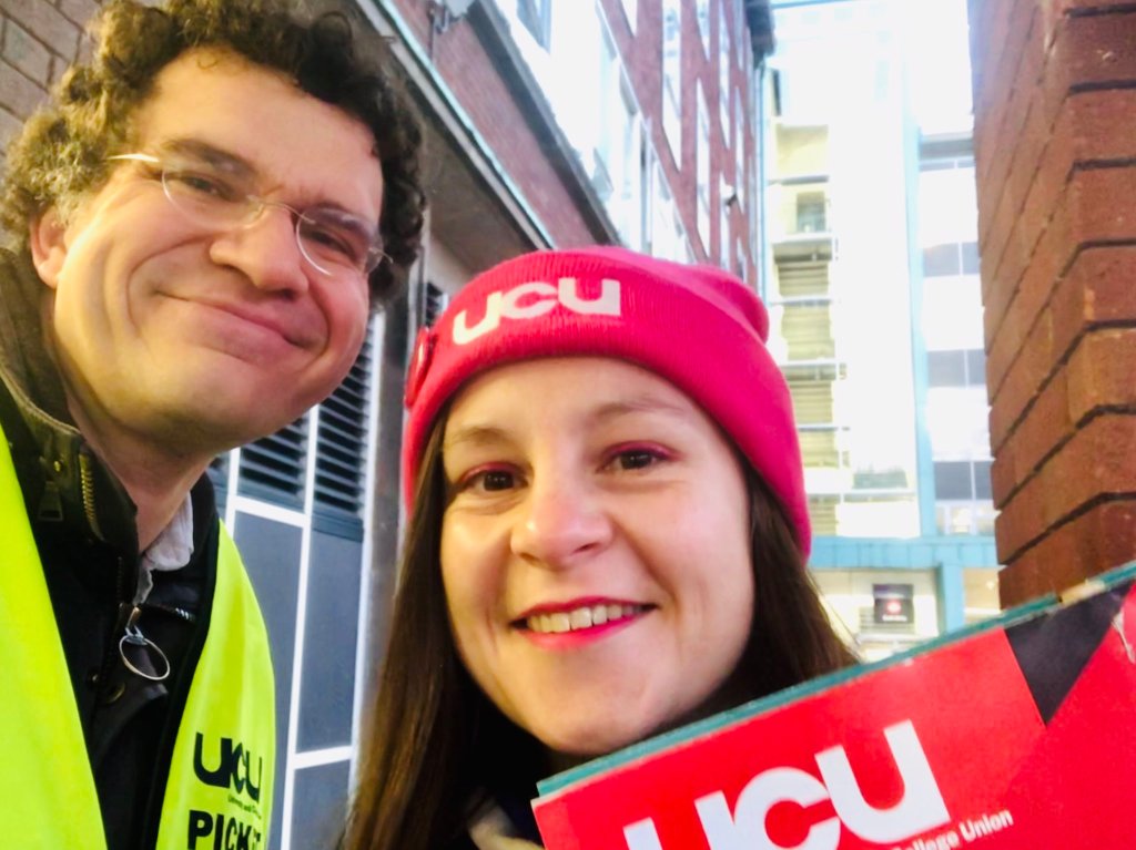 James and Vicky stood between two red brick buildings in Manchester, James (left) has a hi vis yellow UCU Picket vest on, and Vicky is wearing a pink UCU beanie and pink eyeliner. Both are smiling and the top of a UCU placard is visible at the bottom of the shot. 