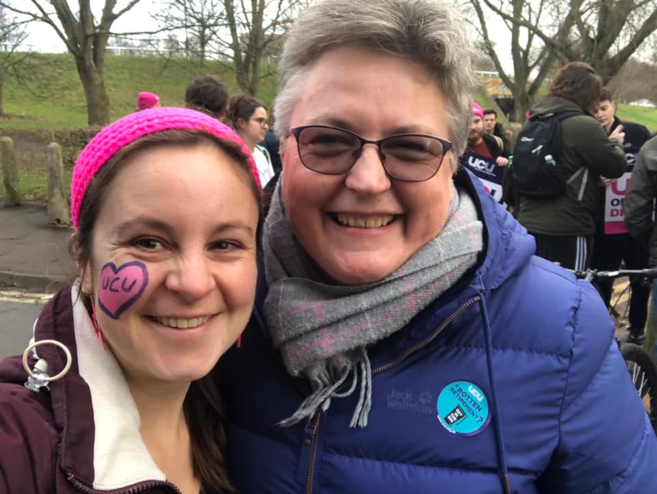 Vicky (on left) and Rachel smiling together in their winter coats on a UCU York Uni picket line. Strikers with placards and posters visible behind them. Rachel wears a USS pensions campaign sticker on her coat and Vicky has a pink and purple UCU heart face paint design on her cheek.